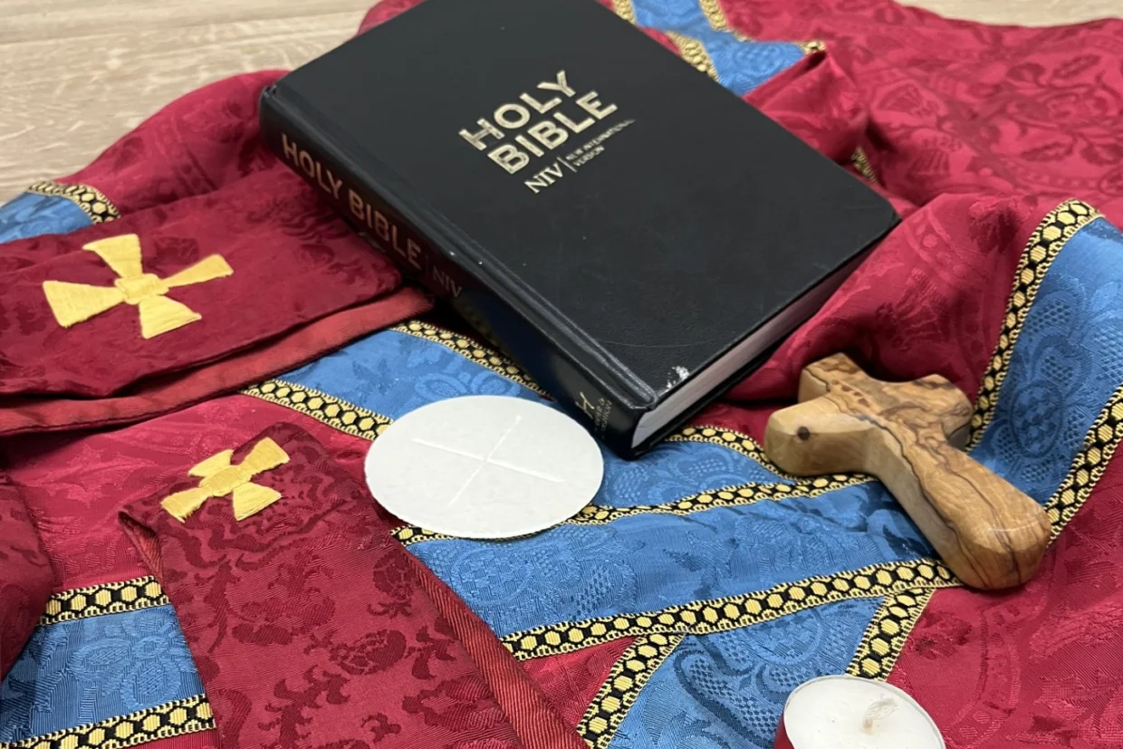 This image shows a bible, a cross, a candle, a communion wafer, a red chasuble and a stole.